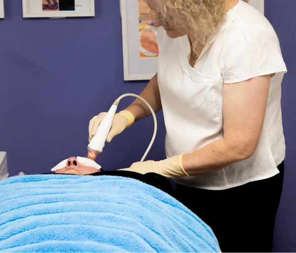 A woman in a white shirt and yellow gloves applies a treatment wand to a person's skin during a skincare or cosmetic procedure, with a blue blanket covering the client and framed skin anatomy posters on a purple wall.