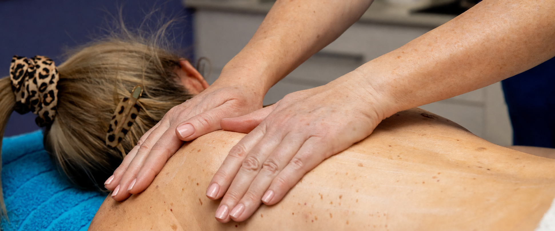 A close-up of a person receiving a massage, lying on their side with another person's hands applying pressure to their upper back. The scene is cropped tightly to show the hands, arms, hair, and bare back.